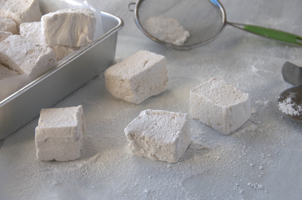 closeup of four marshmallows with Honey Vanilla Marshmallows piled in a parchment lined silver cake pan to the left side and sifter with powdered sugar behind it