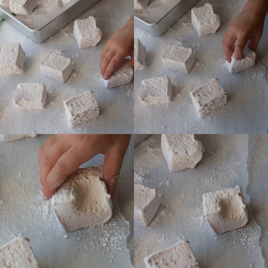 marshmallows being covered in powdered sugar on parchment paper, one marshmallow with a bite taken