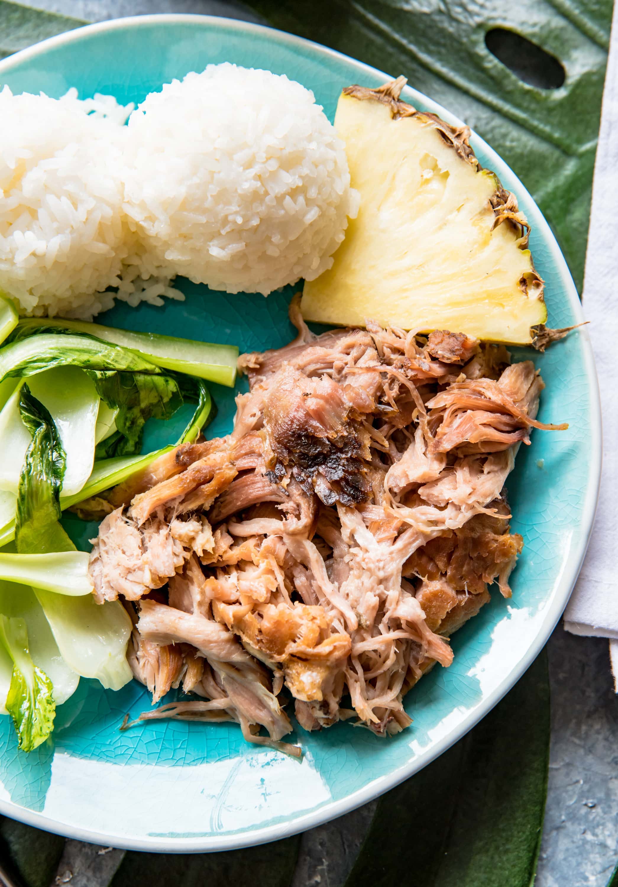 aqua plate with a Hawaiian kalua pork, baby bok choy, pineapple wedge, two scoops of white sticky rice.