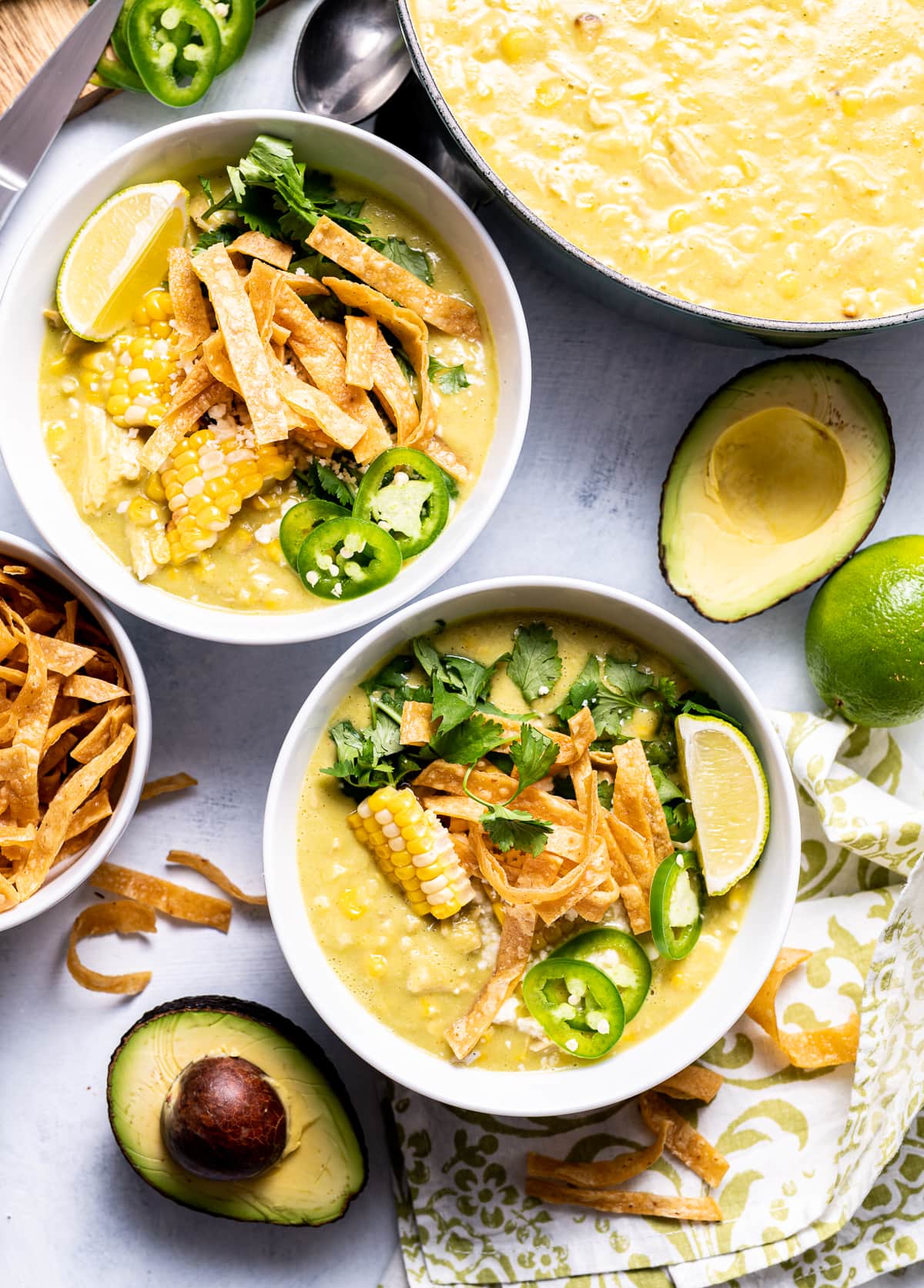 white bowls of green yellow chicken avocado chowder with corn tortilla strips limes jalapeno slices and avocado halves
