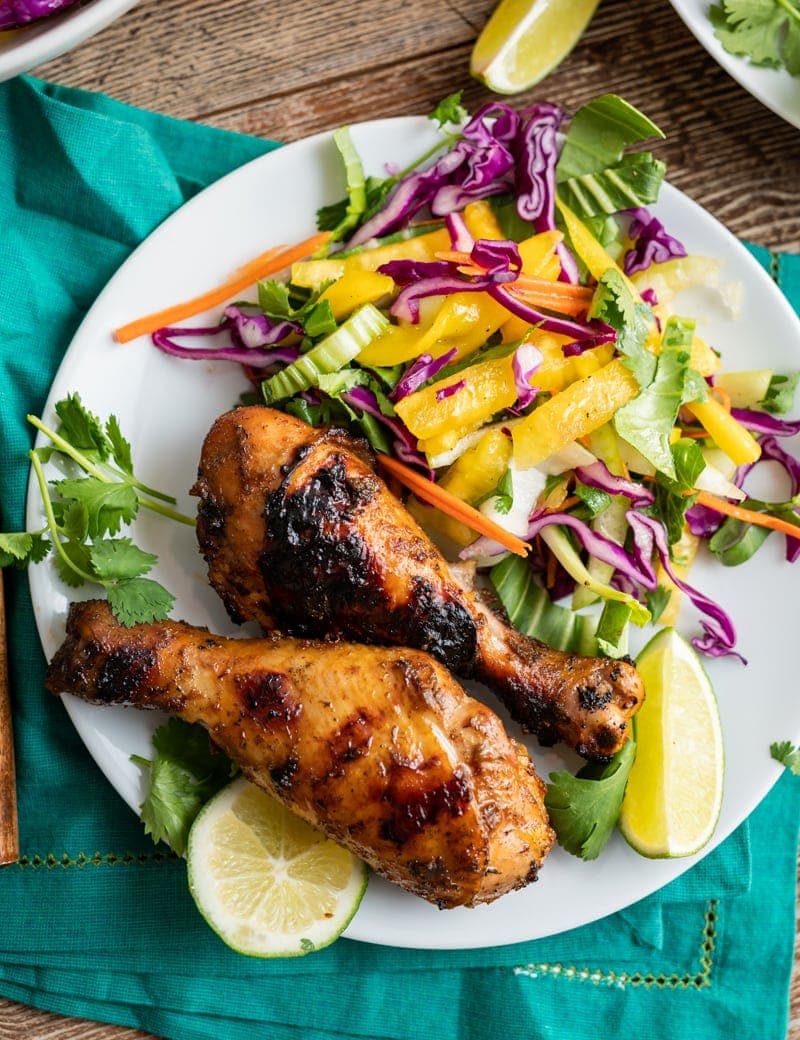close up image of two seasoned grilled chicken drumsticks on a white plate, some mango and bok choy slaw, lime slices, aqua napkin