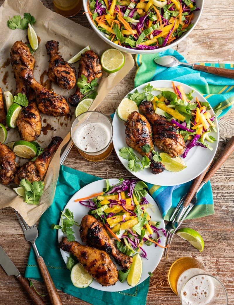 Chicken drumsticks with seasonings and lime slices on a paper lined tray, lime slices, two glasses of beer, two white plates with chicken drumsticks and tropical slaw on them, forks and striped napkins
