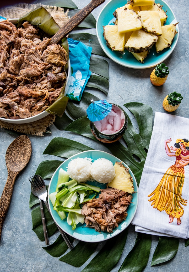 large bowl with shredded kalua pork, small blue plate with serving of kalua pork, scoops of white sticky rice, pineapple wedges, baby bok choy, napkin with hula girl, pineapple salt and pepper shakers.