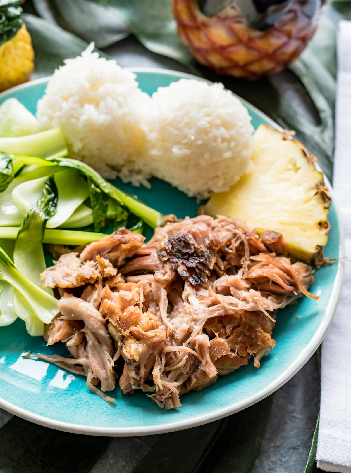 kālua pork on a blue plate with baby bok choy pineapple wedge scoops of white rice