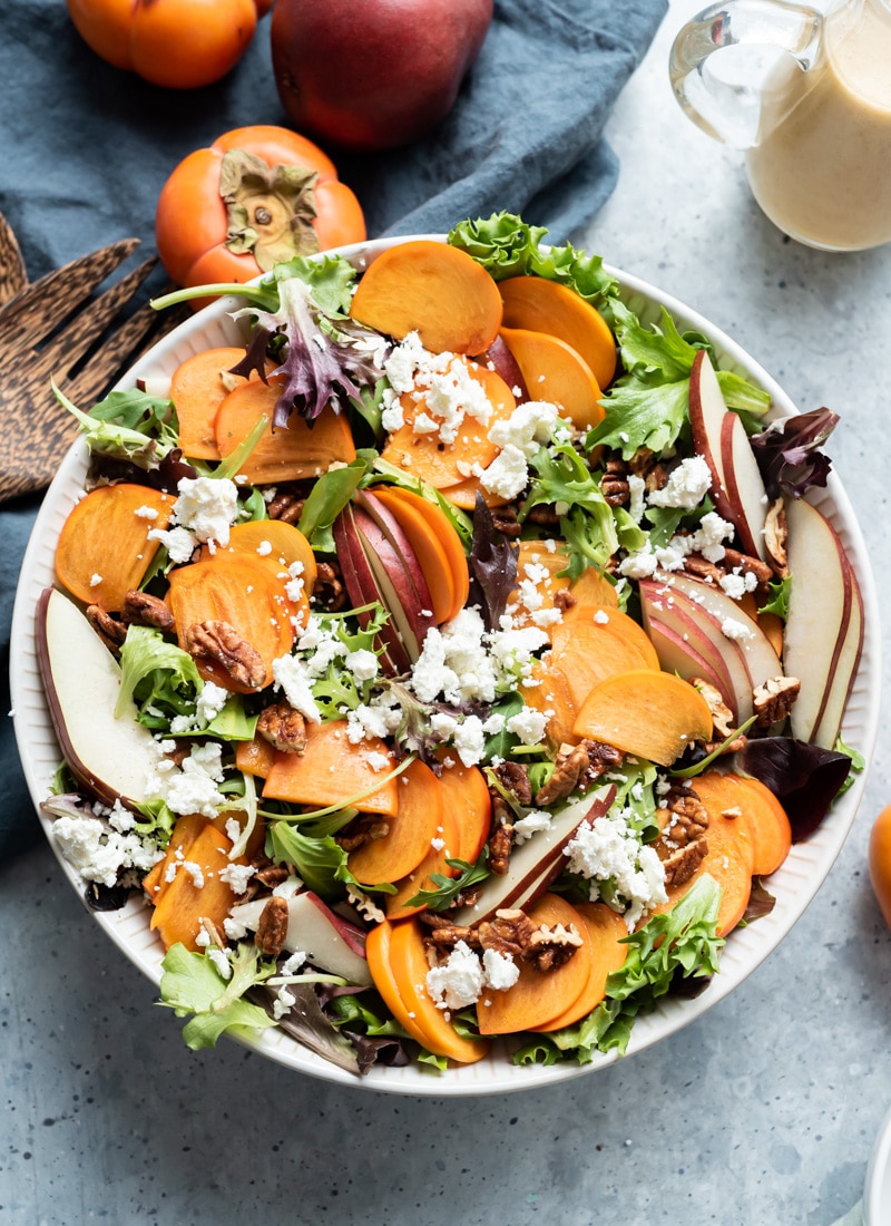 white bowl filled with lettuce, persimmon slices, pear slices, pecans, goat cheese, bottle of vinaigrette