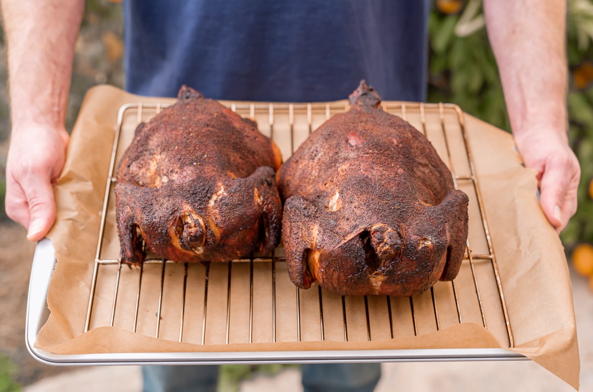 man holding tray of two smoked chickens on a rack on top of parchment paper