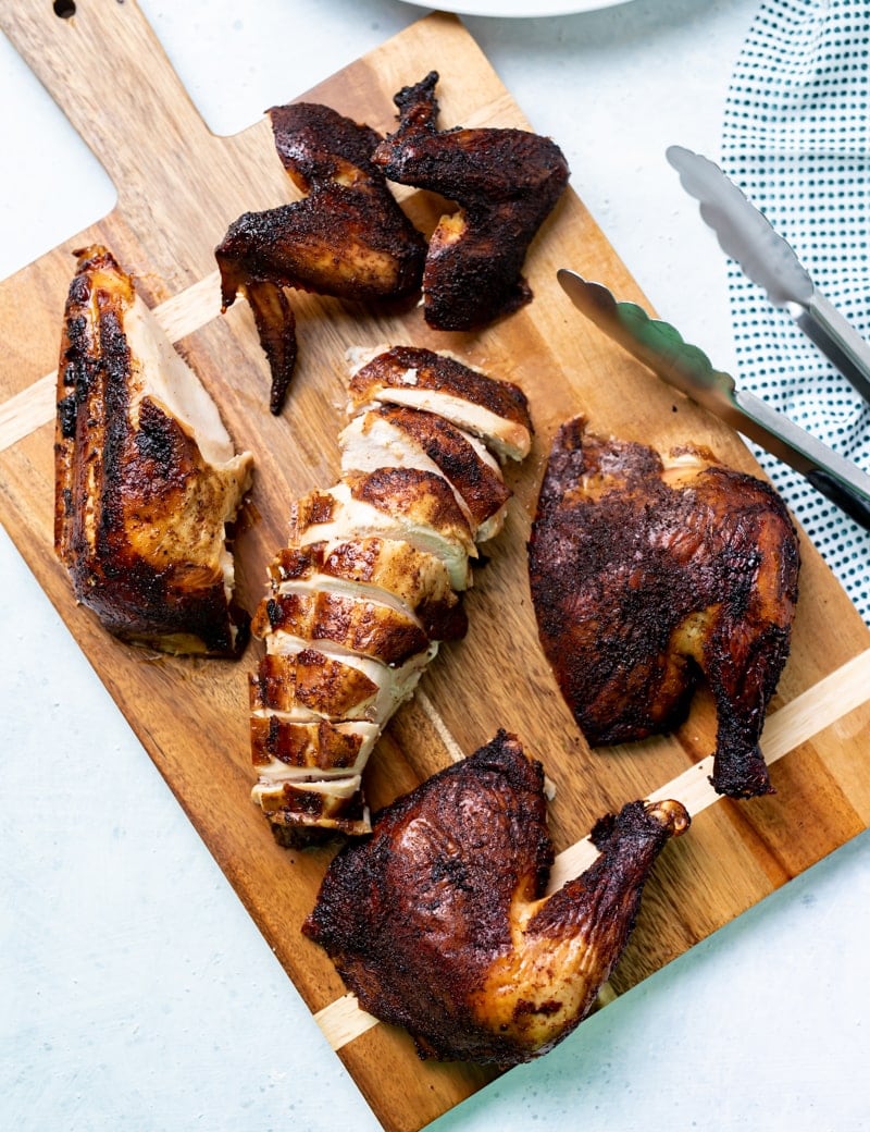 wooden cutting board with handle with various pieces of smoked chicken on it, one piece is sliced, bbq tongs