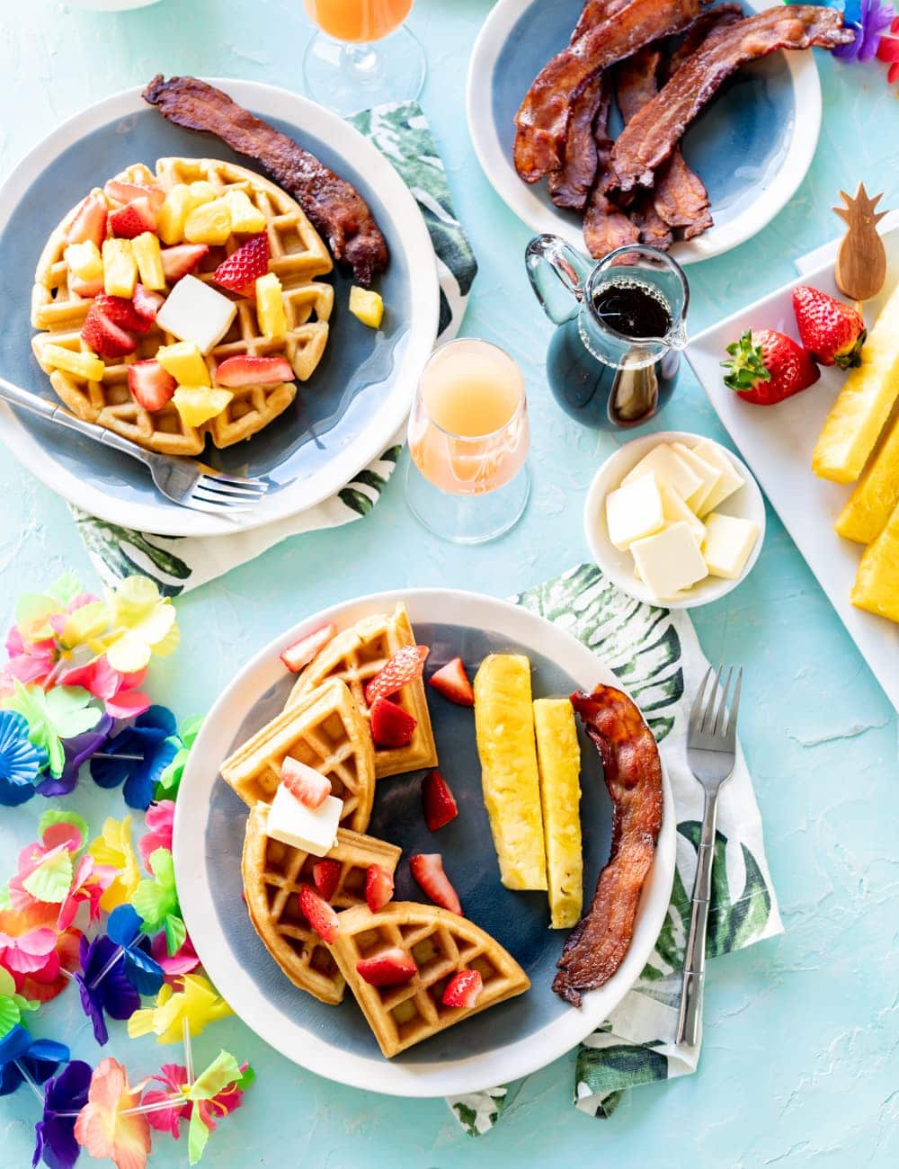 coconut mochi waffles on blue plates with pineapple spears, pieces of cooked bacon, glass with mimosa cocktail in it, glass syrup bottle, rainbow Hawaiian lei on table