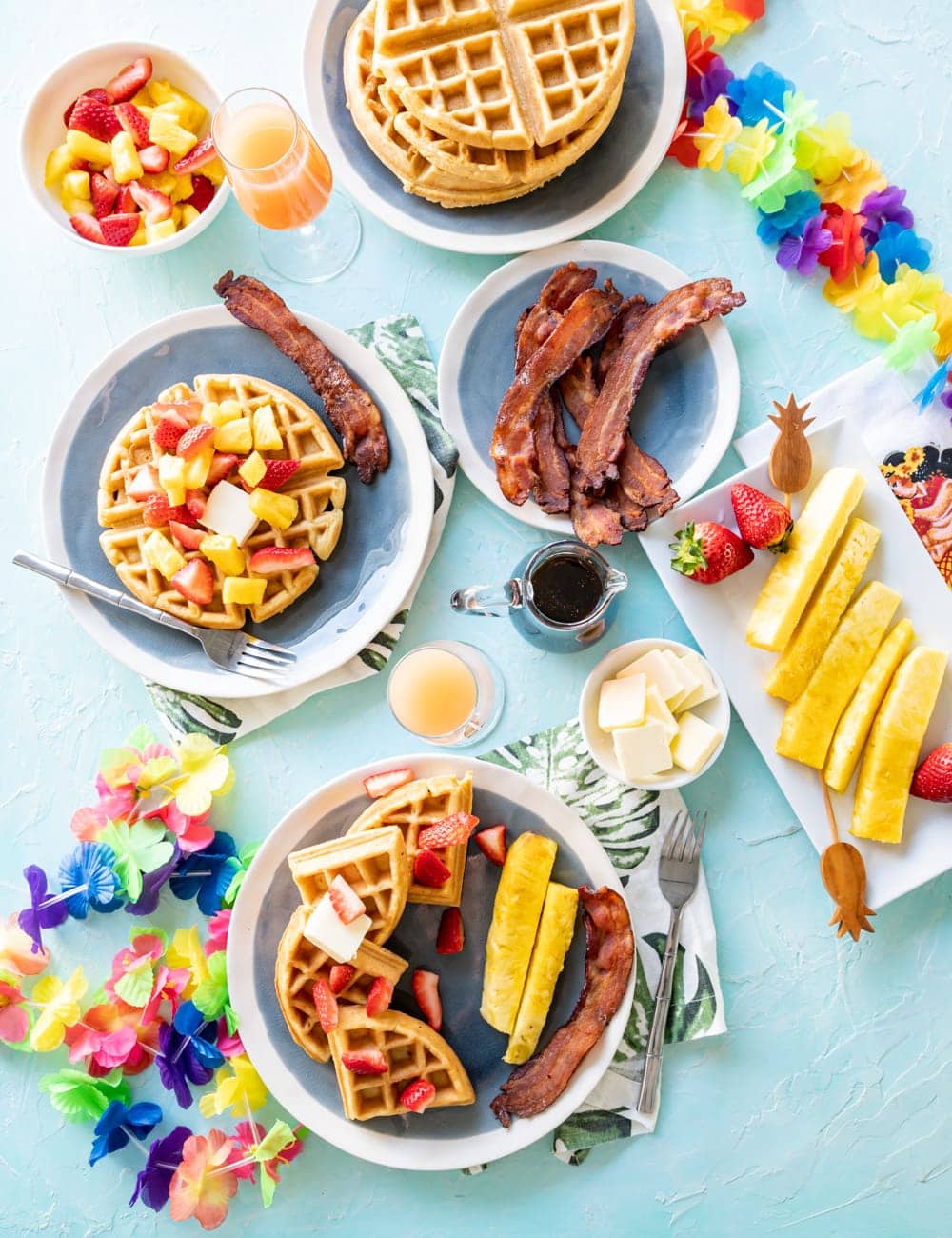 coconut mochi waffles on blue plates with pineapple spears, pieces of cooked bacon, glass with mimosa cocktail in it, glass syrup bottle, rainbow Hawaiian lei on table