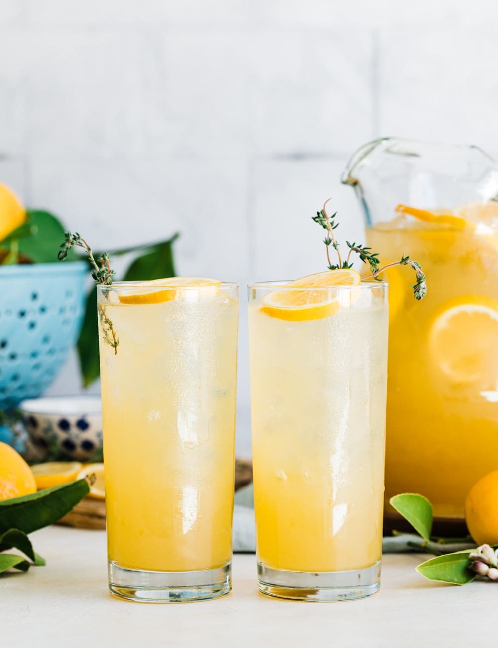 dvě vysoké collins sklenice naplněné s limonádou, plátky citronu a čerstvým tymiánem v sklenice, džbán nebo limonády, čerstvé celé citrony, modrá mísu s citrony, prkénko s plátky citronu