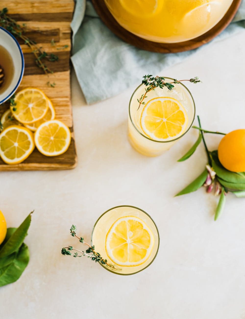  deux grands verres collins remplis de limonade, de tranches de citron et de thym frais dans des verres, pichet ou limonade, citrons entiers frais, bol bleu aux citrons, planche à découper avec des tranches de citron 