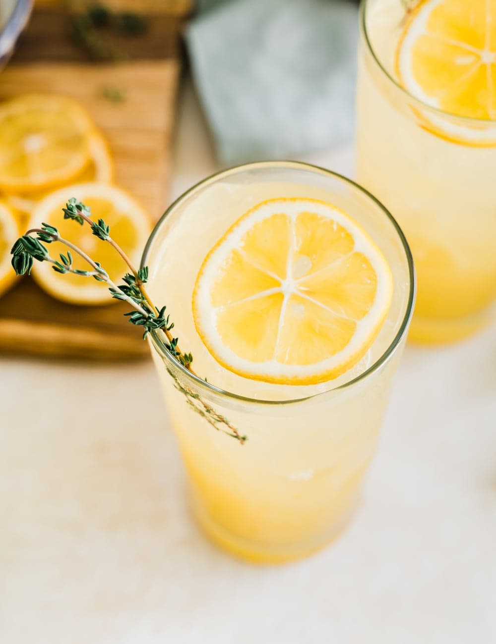 two tall collins glasses filled with lemonade, lemon slices and fresh thyme in glasses, pitcher or lemonade, fresh whole lemons, blue bowl with lemons, cutting board with lemon slices