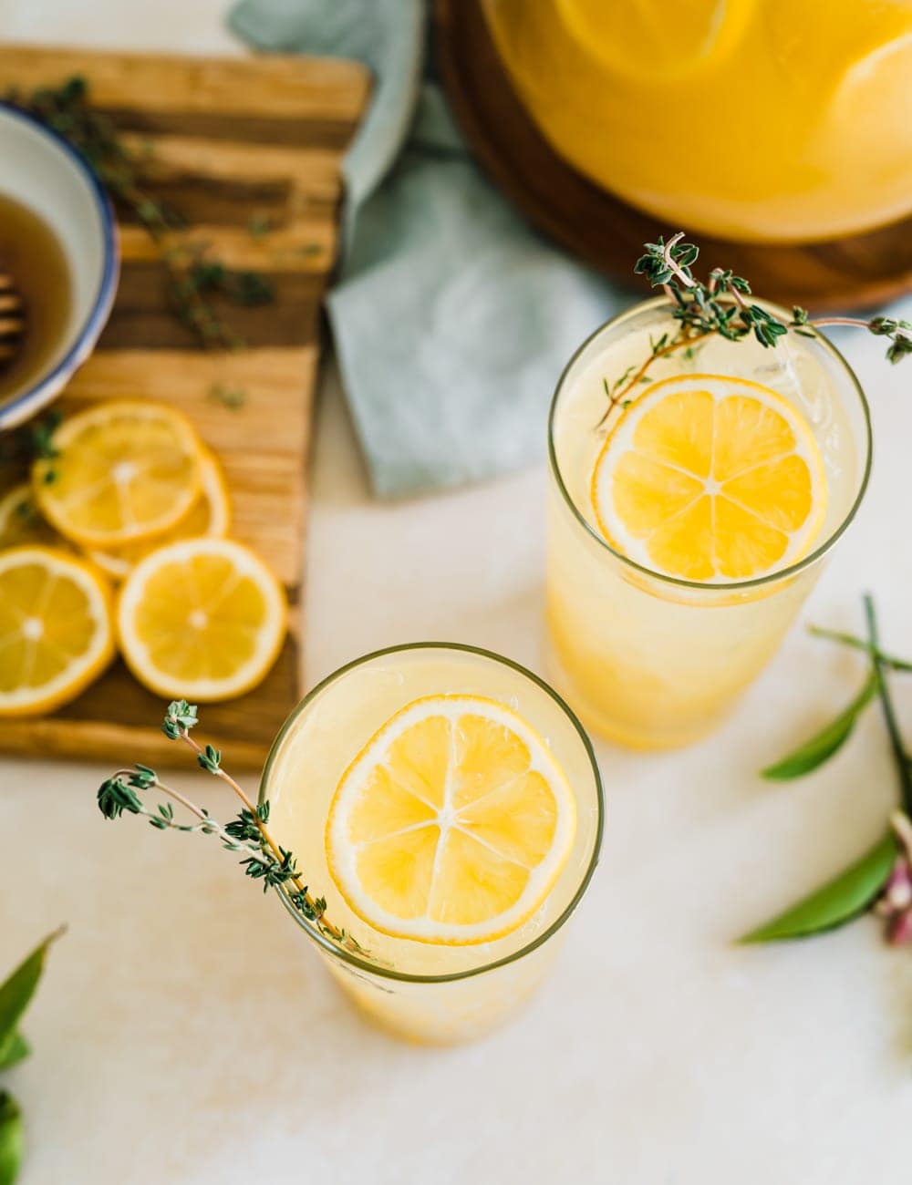 two tall collins glasses filled with lemonade, lemon slices and fresh thyme in glasses, pitcher or lemonade, fresh whole lemons, blue bowl with lemons, cutting board with lemon slices