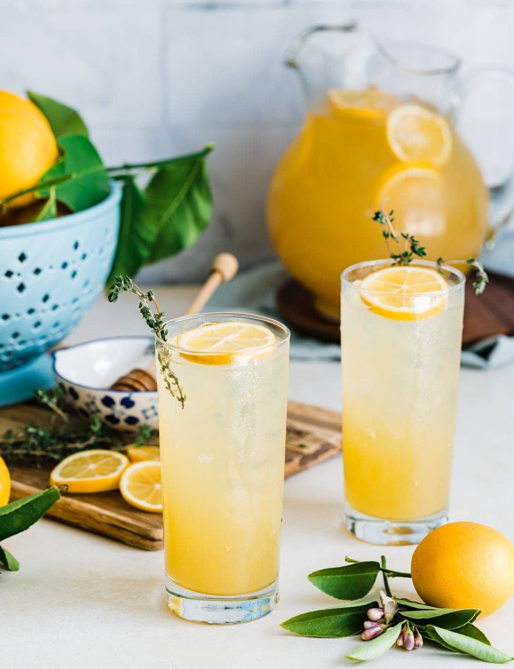 two tall collins glasses filled with lemonade, lemon slices and fresh thyme in glasses, pitcher or lemonade, fresh whole lemons, blue bowl with lemons, cutting board with lemon slices