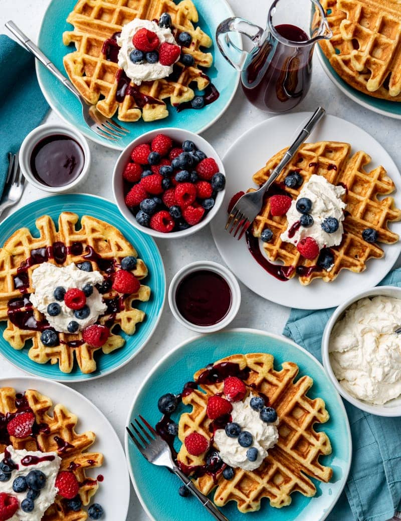 Several white and aqua plates with waffles on each plate, each waffle topped with whipped cream, blueberries and raspberries, dark red syrup, bowl of fresh raspberries and blueberries