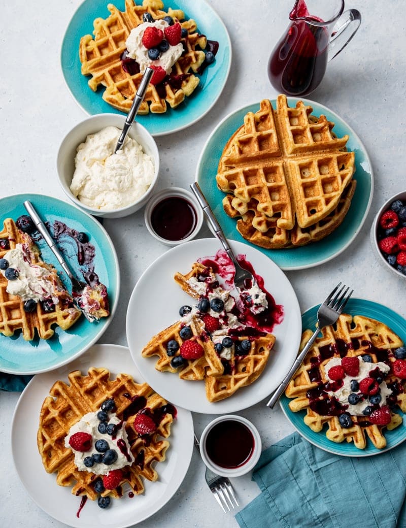 Several white and aqua plates with waffles on each plate, each waffle topped with whipped cream, blueberries and raspberries, dark red syrup, bowl of fresh raspberries and blueberries