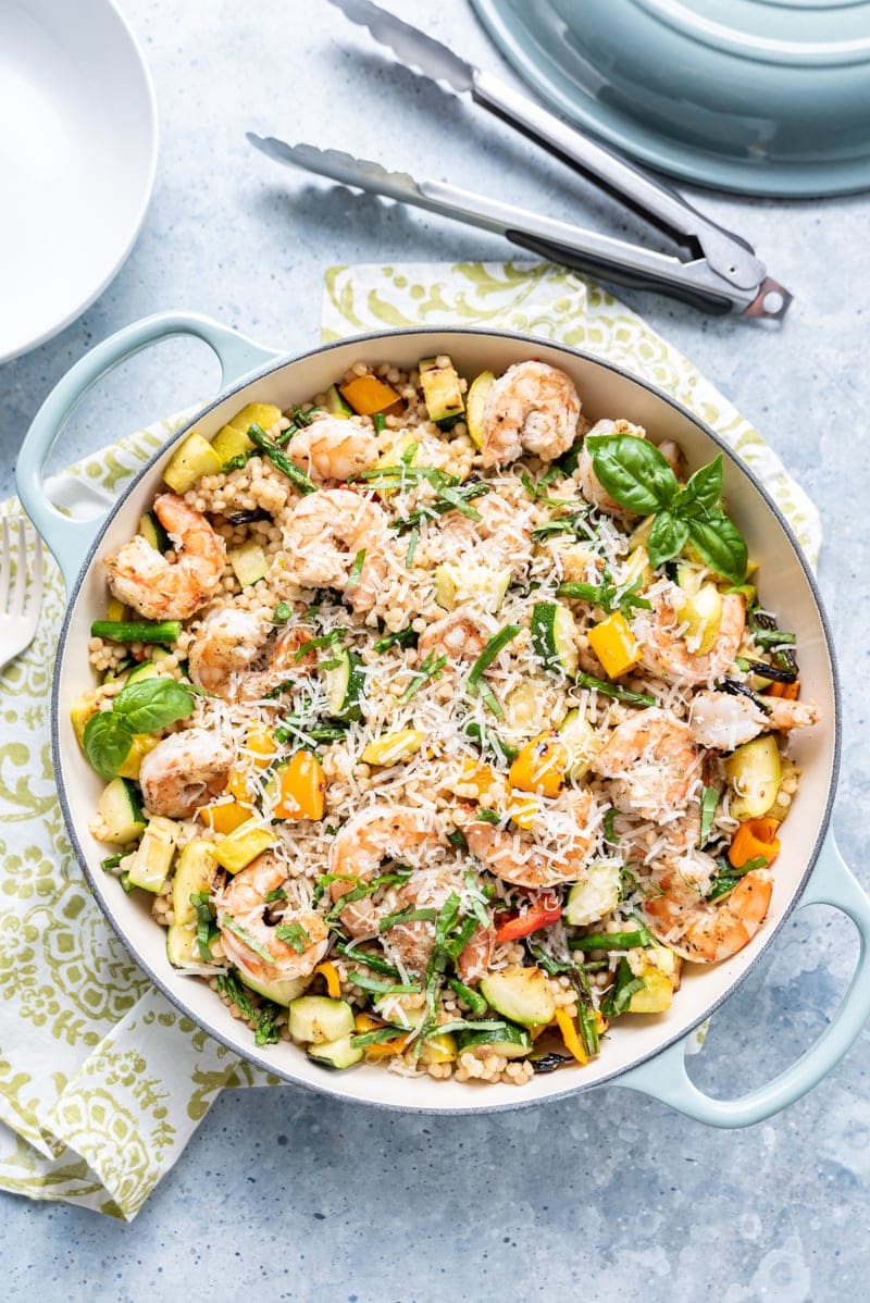 large light blue pan filled with Israeli couscous, cooked shrimp, various squash and peppers, fresh basil, shredded parmesan, metal tongs, green and white napkin