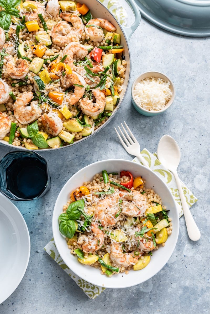 large light blue pan filled with Israeli couscous, cooked shrimp, various squash and peppers, fresh basil, shredded parmesan, small white bowl filled with same ingredients, green and white napkin