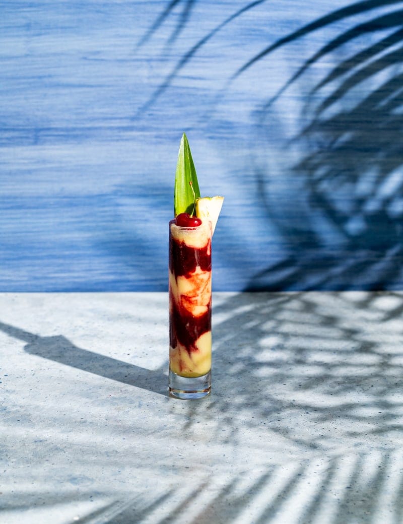 tall skinny glass with alternating layers of yellow and red frozen cocktail, cherry and pineapple leaf in glass