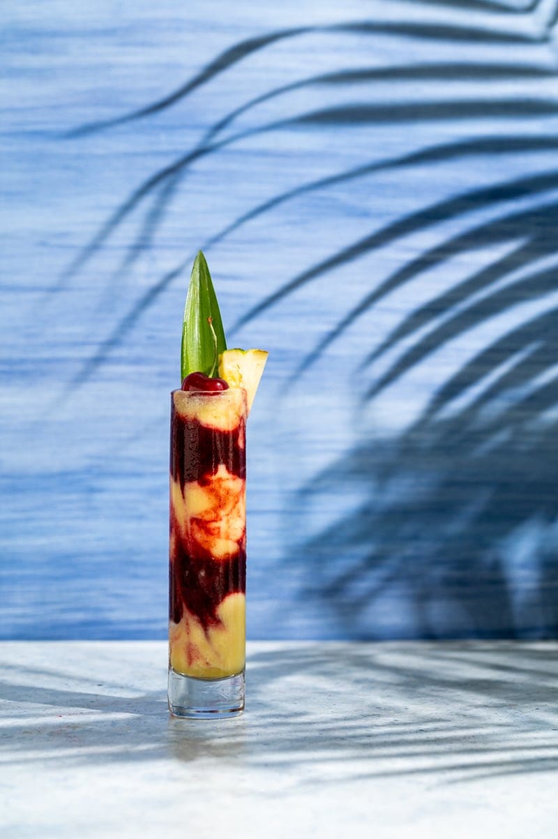 tall skinny glass with alternating layers of yellow and red frozen cocktail, cherry and pineapple leaf in glass