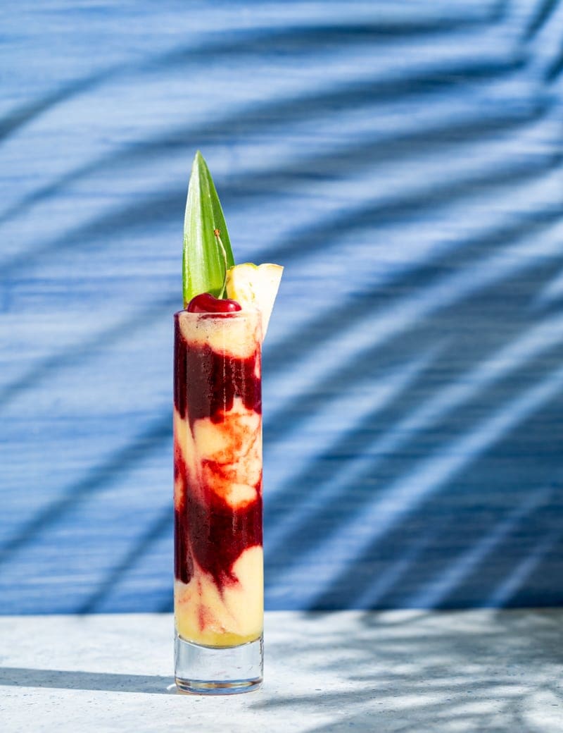 tall skinny glass with alternating layers of yellow and red frozen cocktail, cherry and pineapple leaf in glass