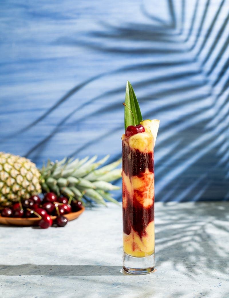 tall skinny glass with alternating layers of yellow and red frozen cocktail, cherries, palm leaf shadow