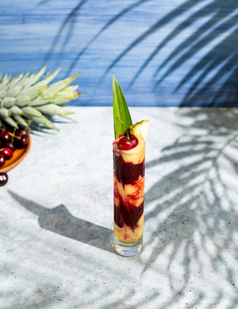 tall skinny glass with alternating layers of yellow and red frozen cocktail, cherries, palm leaf shadow