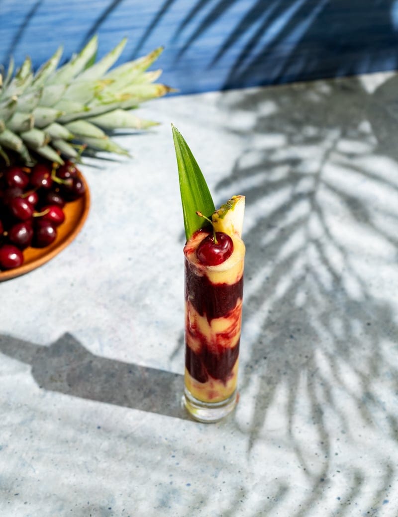 tall skinny glass with alternating layers of yellow and red frozen cocktail, cherries, palm leaf shadow