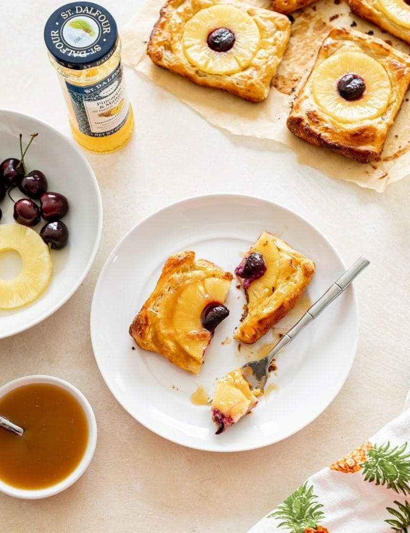 Overhead photo of Pineapple Puff Pastry Tarts with Pineapple Rum Sauce, tarts on small white plates, one cut into pieces with a fork, pineapples and cherries on a white plate, jar of jam, beige background, pineapple towel, rum sauce in small dish www.pineappleandcoconut.com