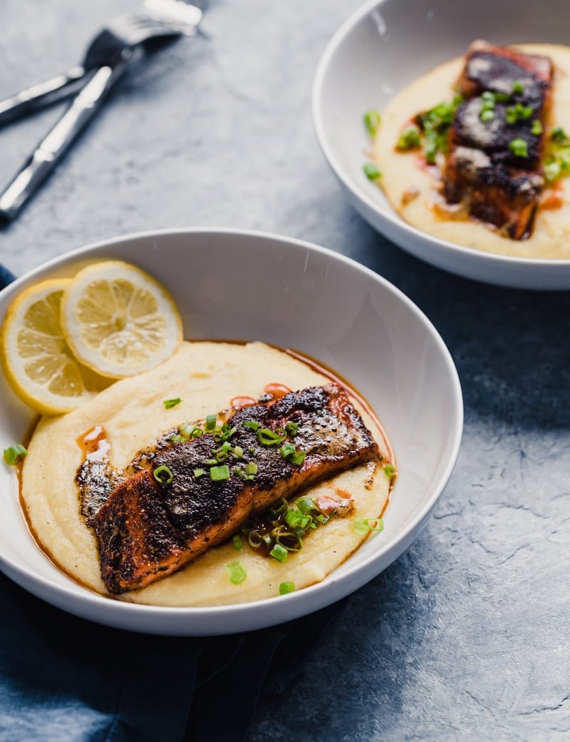 angled photo of two white bowls of wild sockeye salmon fillets served over cheddar grits with hot sauce and lemon slices