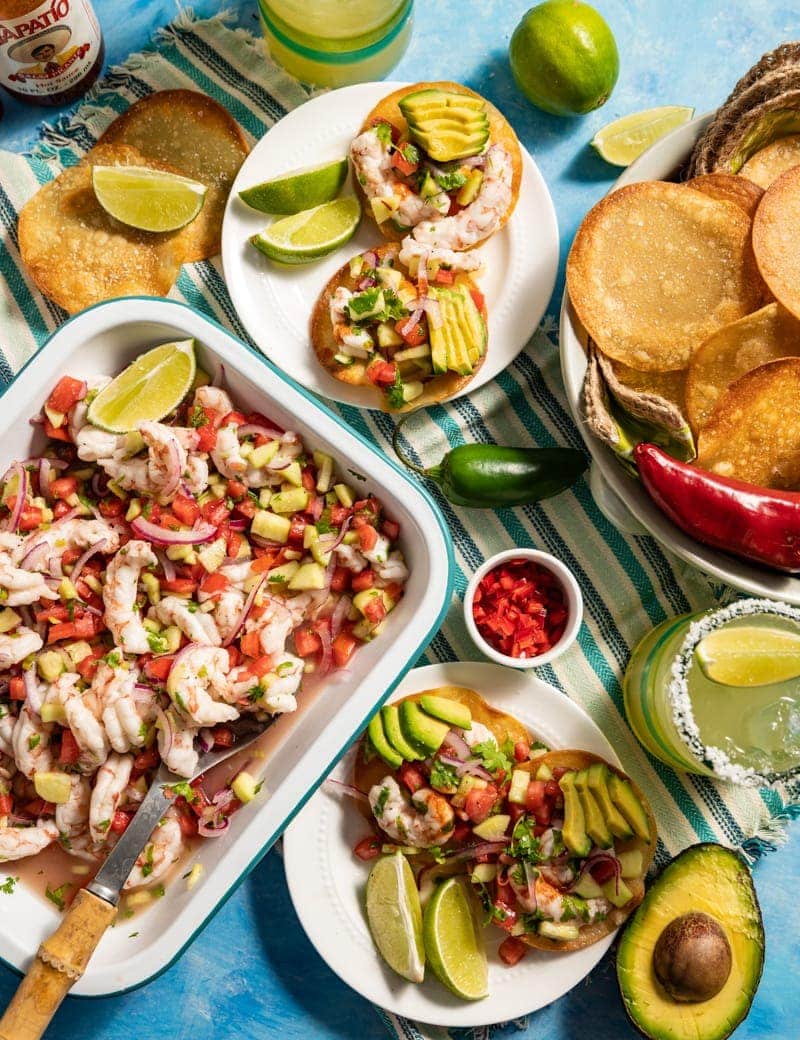 Beautiful food photography of Mini Shrimp Ceviche Tostada recipe with tray of shrimp ceviche, margaritas, lime slices, diced chilies in small bowls basket of round chips small plates with servings of ceviche