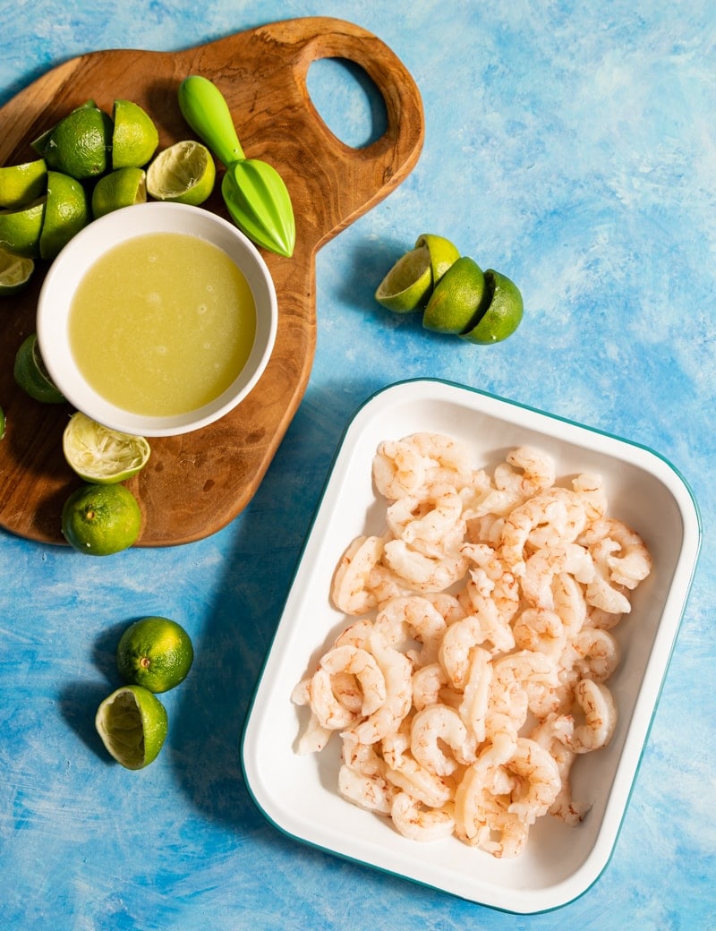 Shrimp Ceviche prep pic, shrimp in a shallow pan, limes cut in half on a wood board, lime juice in a white bowl