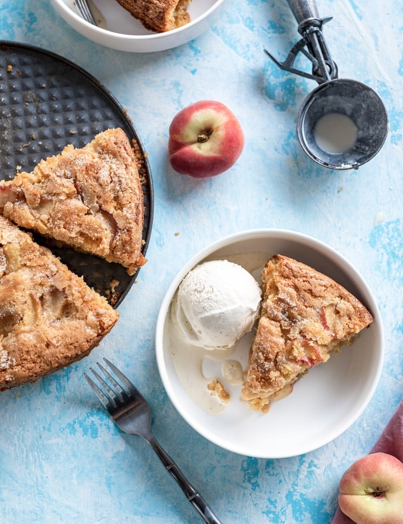 overhead close up of Summertime Brown Sugar White Peach Buttermilk Cake slice in white bowl with scoop of vanilla ice cream, peaches, ice cream scoop, partial view of slices of cake on cake pan www.pineappleandcoconut.com