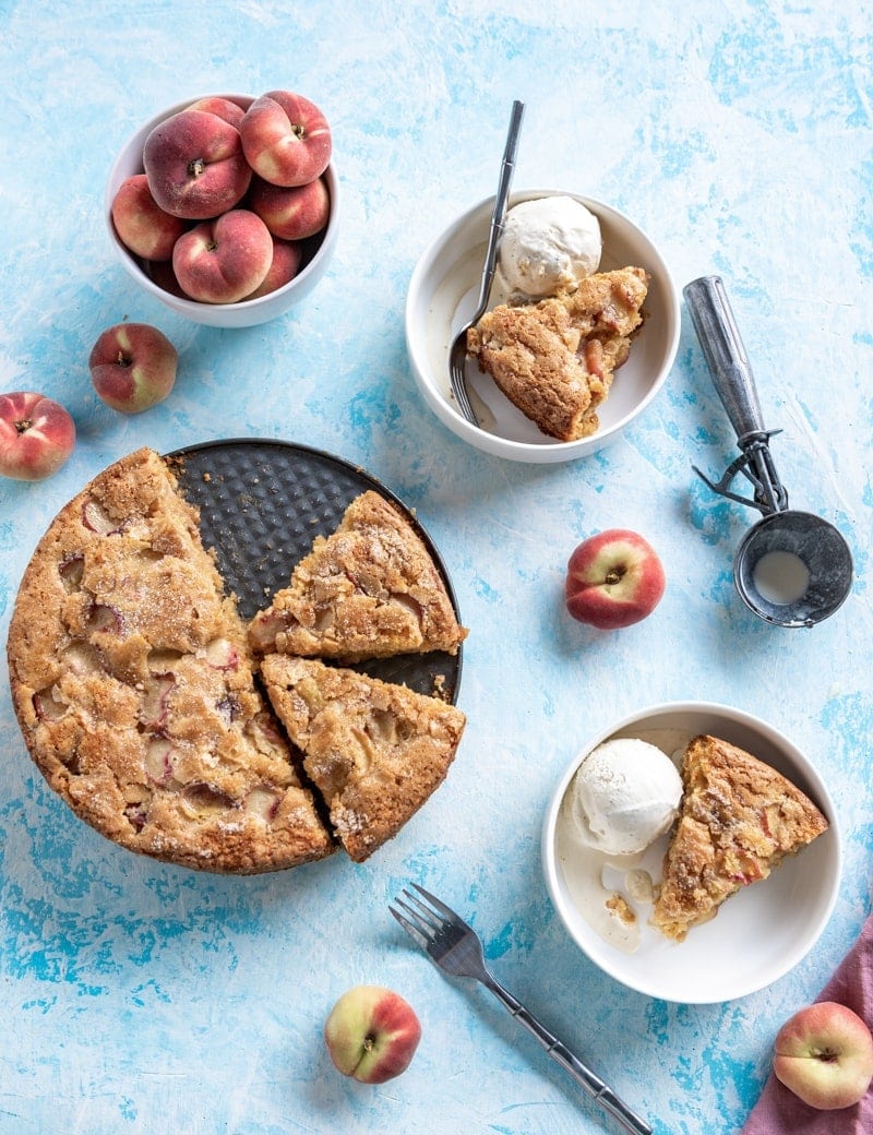 Overhead photo of Summertime Brown Sugar White Peach Buttermilk Cake on baking pan, two white bowls of slices of cake with scoops of ice cream white bowl with peaches, ice cream scoop, blue background www.pineappleandcoconut.com