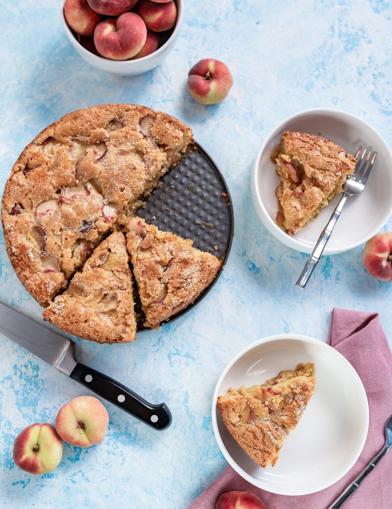 overhead photo of Summertime Brown Sugar White Peach Buttermilk Cake in pan, peaches in white bowl, knife, pink napkin, two white bowls with slices of cake in them, forks www.pineappleandcoconut.com