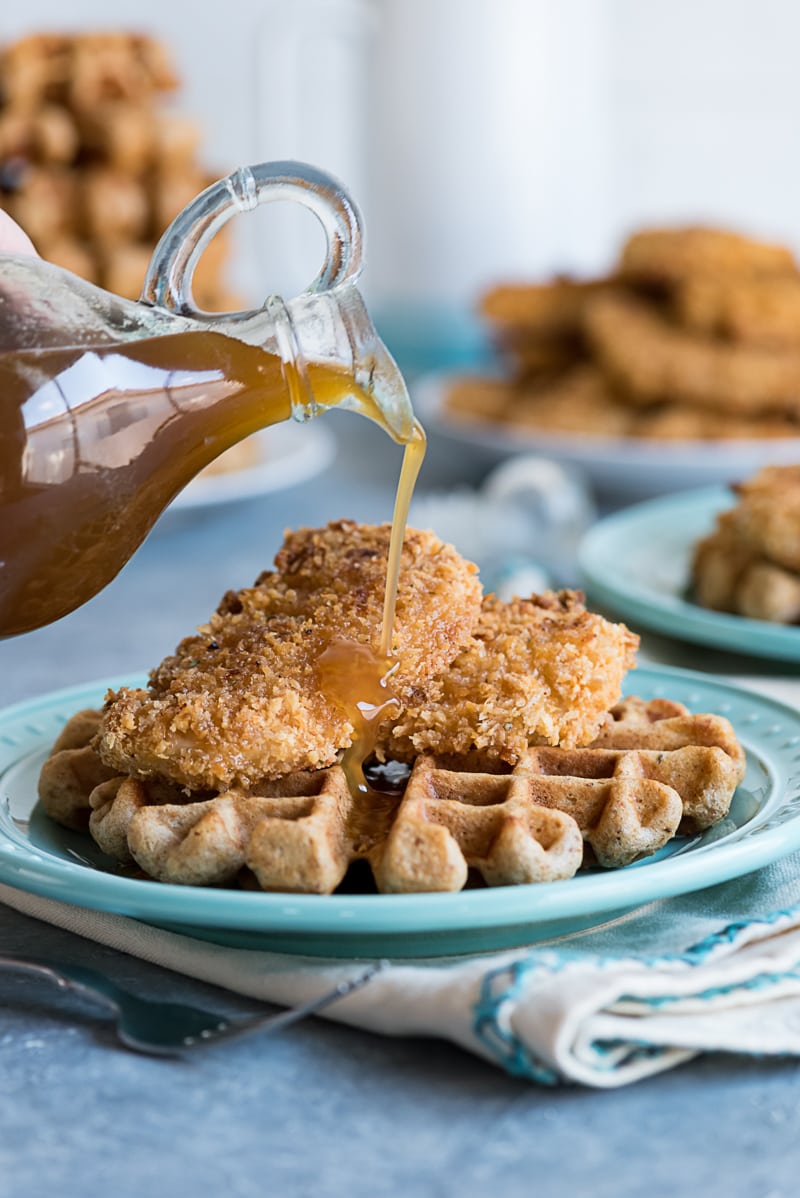 syrup being poured out of a bottle onto a piece of baked chicken on top of a waffle on a blue plate
