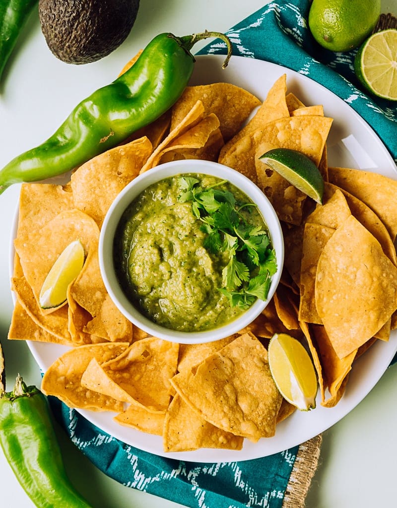 green salsa in a small white bowl on a plate filled with chips, large raw hatch chiles, limes, avocado
