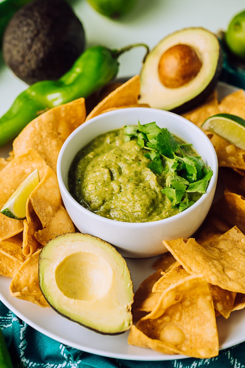 green roasted hatch chile salsa in a small white bowl on a plate filled with chips, large raw green chiles, limes, avocado