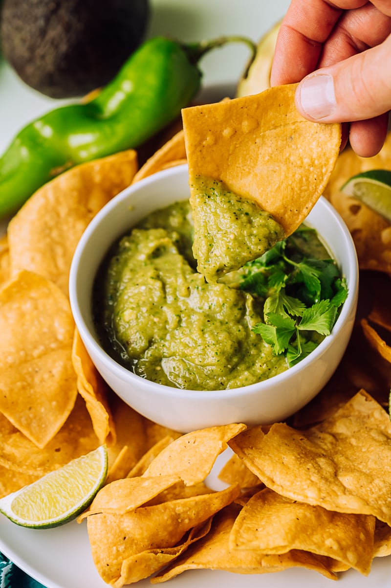 hand holding a chip with some green salsa on it over a small bowl of green salsa, plate of chips, lime slices, raw green chile, avocado