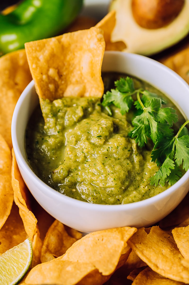 green roasted hatch chile salsa in a small white bowl on a plate filled with chips, large raw green chiles, limes, avocado