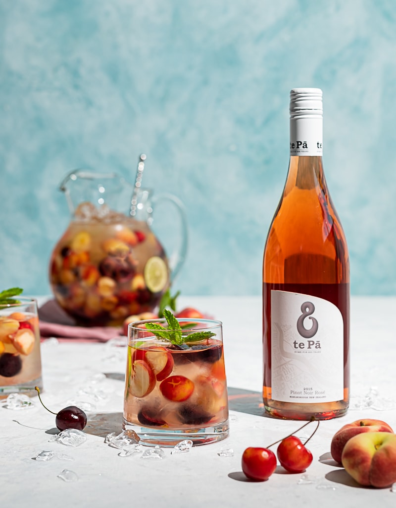 Pitcher filled with cherries and peaches and wine, two glasses filled with ice, wine, fruit, cherries and peaches scattered on table, bottle of te pa rosé wine