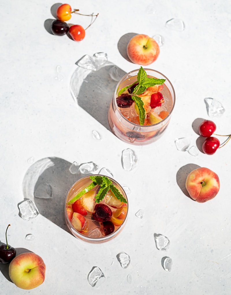 Two glasses filled with ice, wine, fruit, cherries and peaches scattered on table