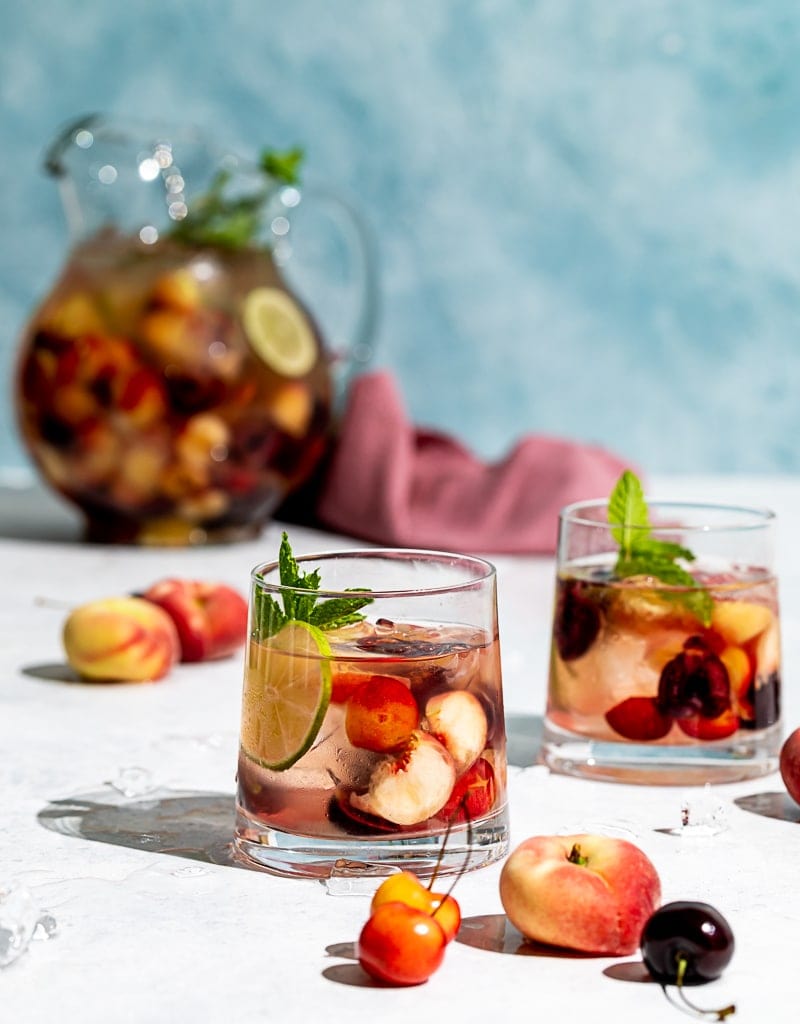 Pitcher filled with cherries and peaches and wine, two glasses filled with ice, wine, fruit, cherries and peaches scattered on table