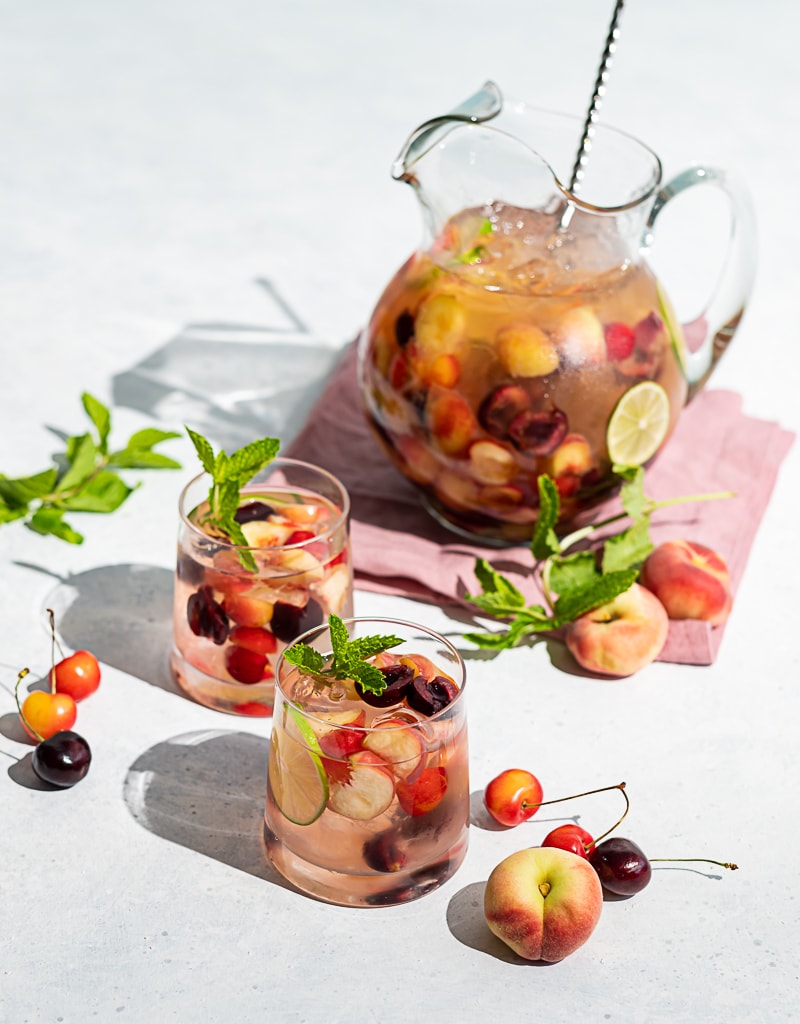 Pitcher filled with cherries and peaches and wine, two glasses filled with ice, wine, fruit, cherries and peaches scattered on table