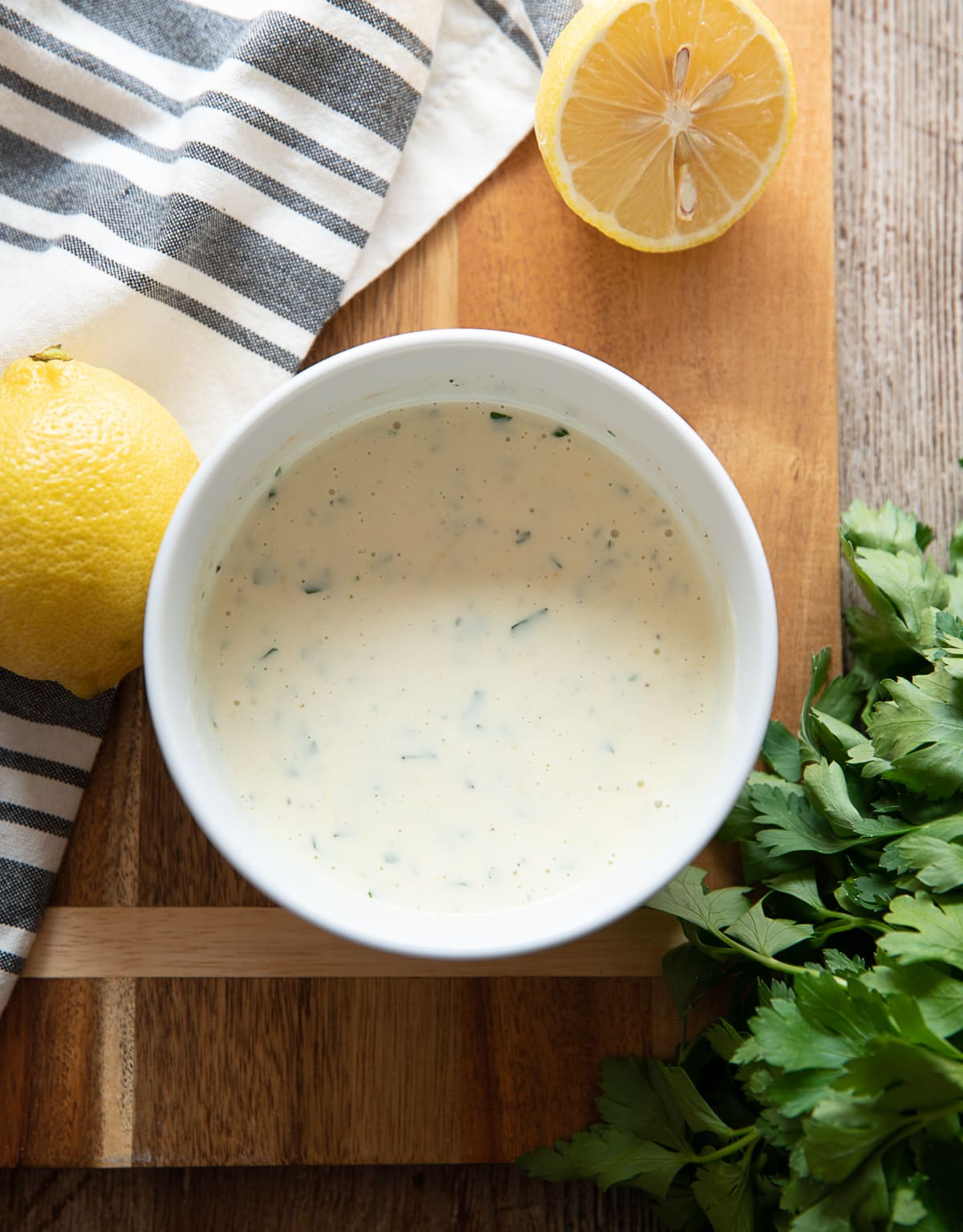white bowl of lemon mayonnaise sauce with parsley, brown cutting board, blue and whits striped napkin , lemons