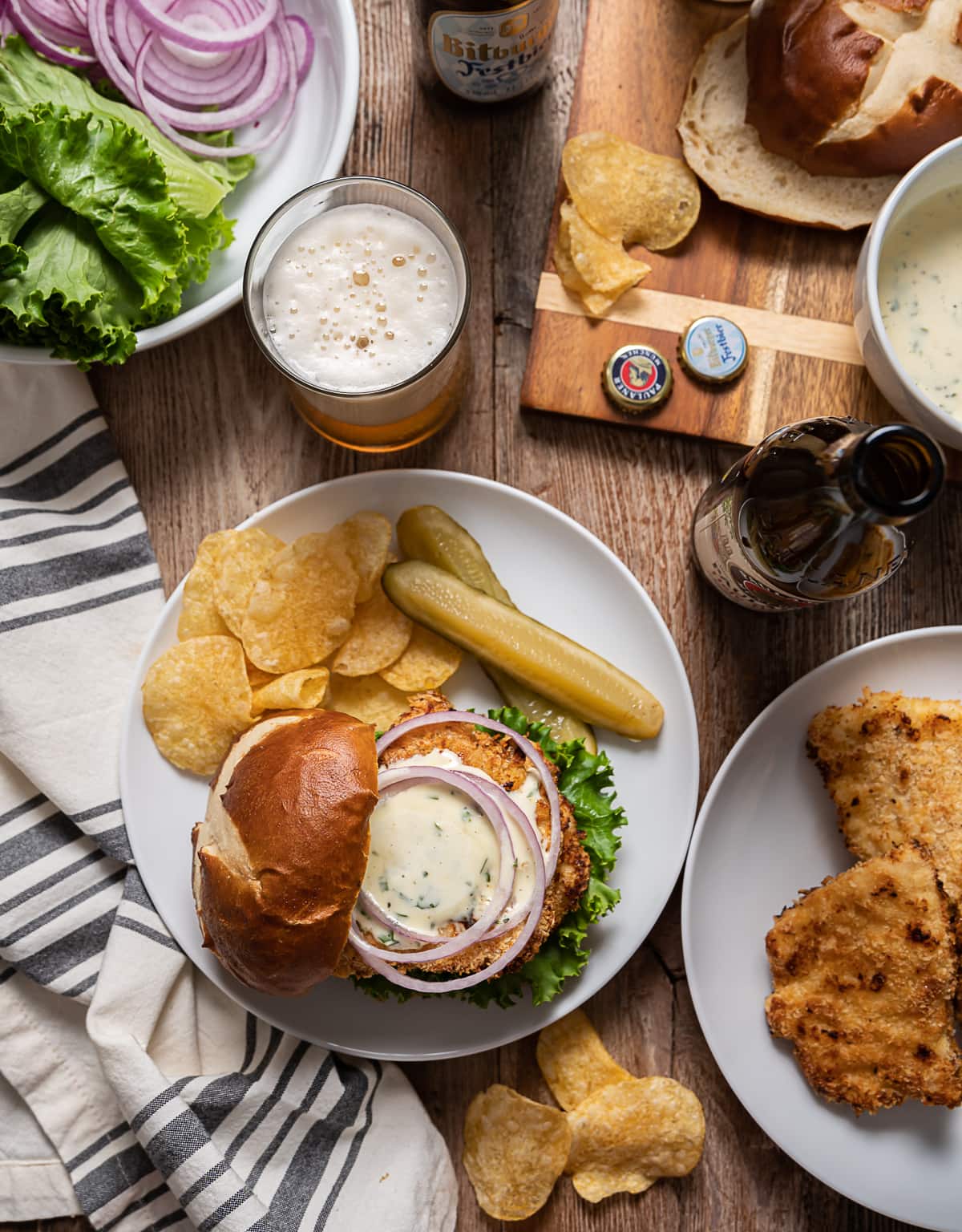 chicken schnitzel sandwich with mayonnaise sauce, red onion rings, plate with thin fried chicken, bottle and glass of beer, bowl with lettuce and onions, cutting board with pretzel buns and bowl of mayonnaise sauce.