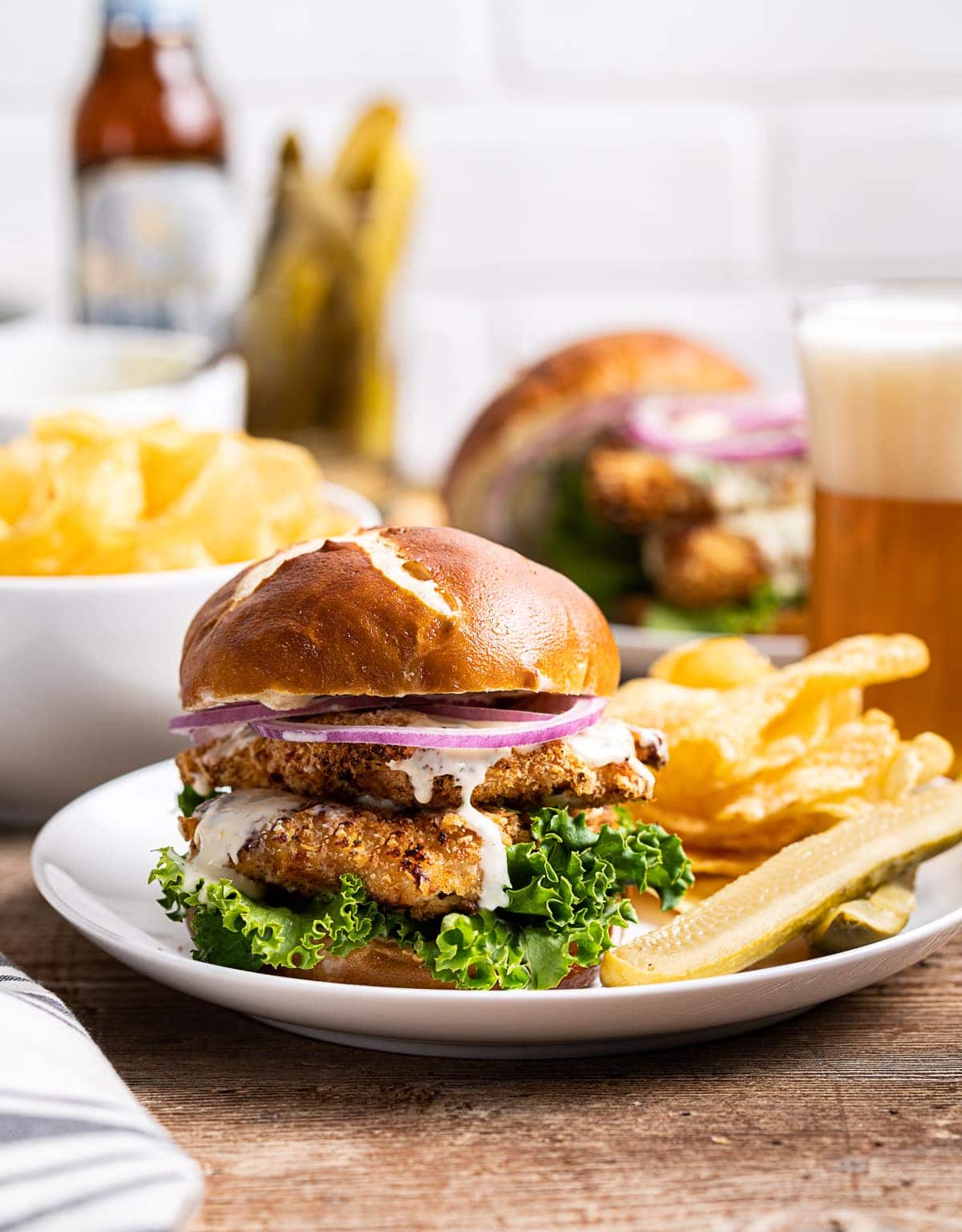 chicken schnitzel sandwiches on pretzel bun with green lettuce red onion kettle chips pickles and glass of beer