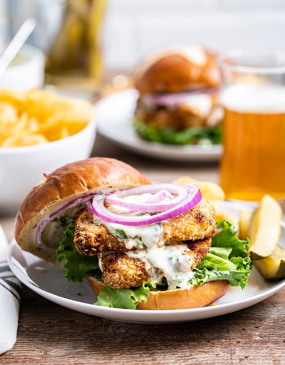 two pieces of breaded air fryer chicken sandwiched in a pretzle bun with green lettuce, red onion slices, pickles and chips on plate, bowl of chips, glass and bottle of beer