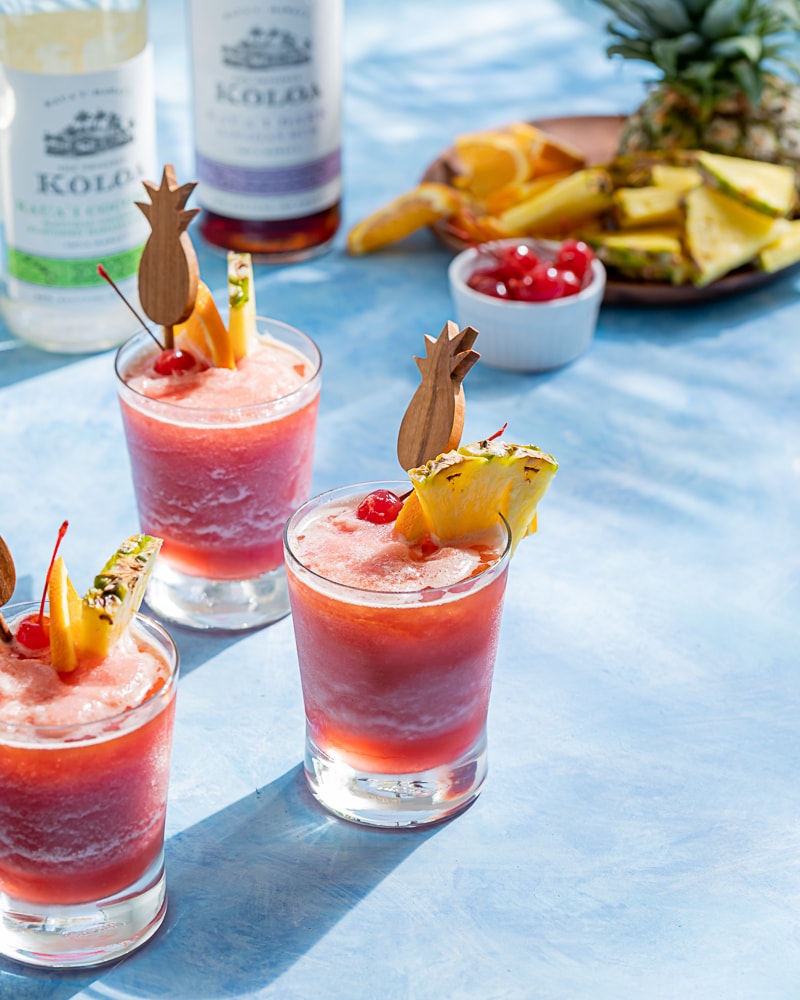 Pink frozen cocktails, pineapple wood cocktail stick, pineapple wedge, orange slice, maraschino cherry, blue background