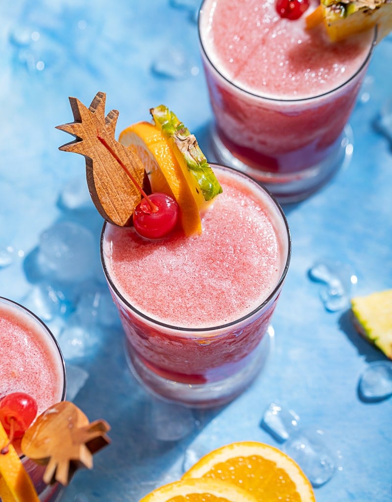 Pink frozen rum runner cocktails, pineapple wood cocktail stick, pineapple wedge, orange slice, maraschino cherry, blue background